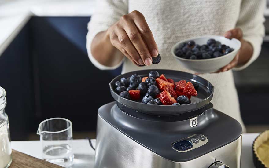 Une personne prépare un dessert sain en ajoutant délicatement des myrtilles à des fraises dans un plat posé sur un appareil de cuisine moderne.