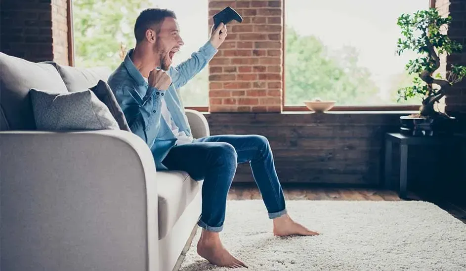 A smiling man dressed in denim is sitting on a grey sofa, holding up a controller in a modern living room with a brick wall.