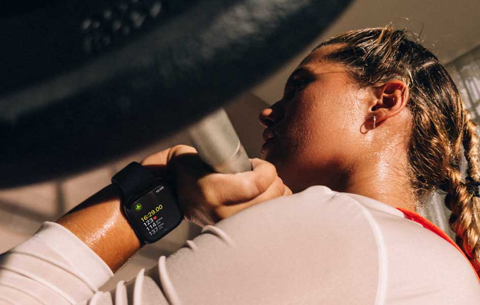 Een sporter traint intensief met een halterstang. Ze draagt een zwarte smartwatch en een licht shirt, terwijl zonlicht haar gezicht en schouders verlicht.
