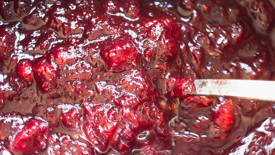 Een close-up van rode confituur met stukjes fruit en een zilveren lepel die erdoor roert.