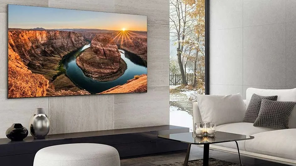 A Samsung TV is mounted on the wall, displaying a stunning canyon river at sunset, in a modern, bright living room with minimalist decor.