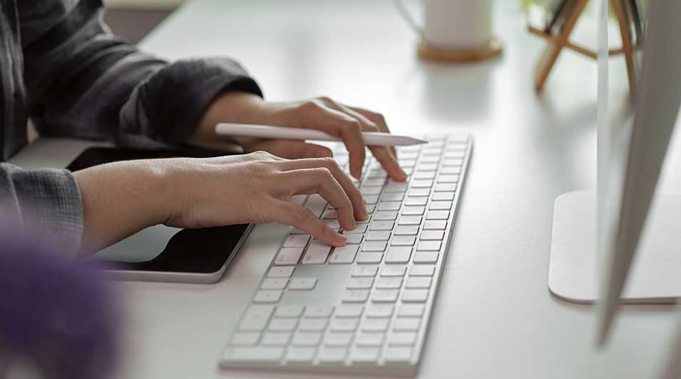 Mains tapant sur un clavier blanc. Un stylet blanc repose sur le clavier. Une tablette noire est à gauche. Bureau blanc.