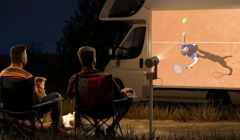 Two men sit outside at night in chairs, watching a tennis match projected onto a camper by a projector. No brand is visible.