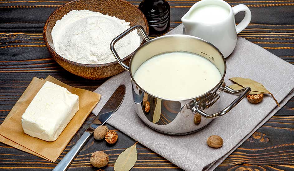 Op een houten tafel staan ingrediënten: bloem in een kom, boter, melk in een kan en een pan met een witte vloeistof.