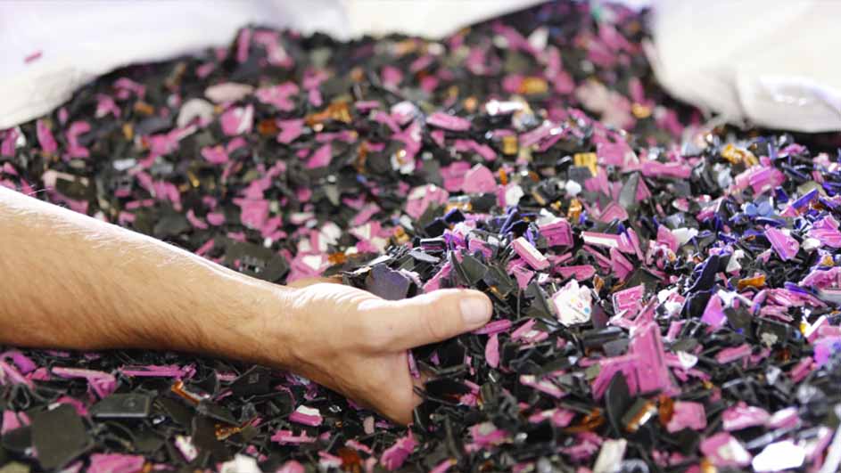 Een hand grijpt in een stapel gebroken, kleine stukjes roze en zwart plastic in een witte zak.