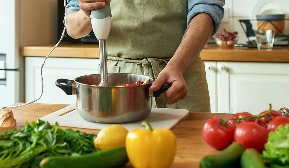 Een roestvrijstalen pan met rode groenten wordt gemixt met een staafmixer, omringd door groenten op een houten aanrecht.