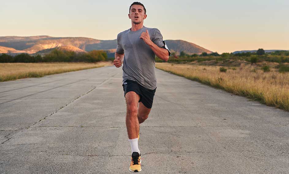 Un homme court sur une route grise, portant un t-shirt gris, un short noir et des chaussures de course orange et noires.