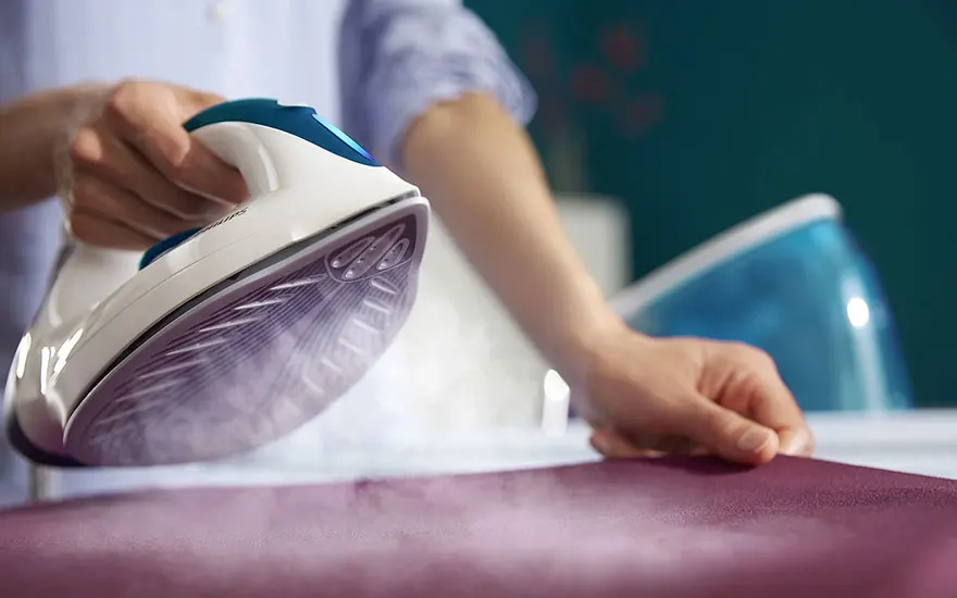 Repassage avec un fer blanc et bleu. La vapeur s'échappe sur un tissu violet. Réservoir d'eau bleu visible.