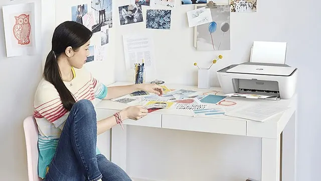 Een witte printer staat op een wit bureau bezaaid met papier en kantoorbenodigdheden. Aan de muur hangen foto's.