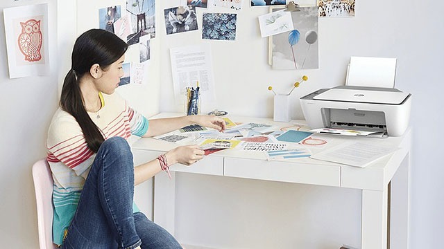 Une imprimante blanche est posée sur un bureau blanc avec des papiers colorés. Des photos sont épinglées au mur.