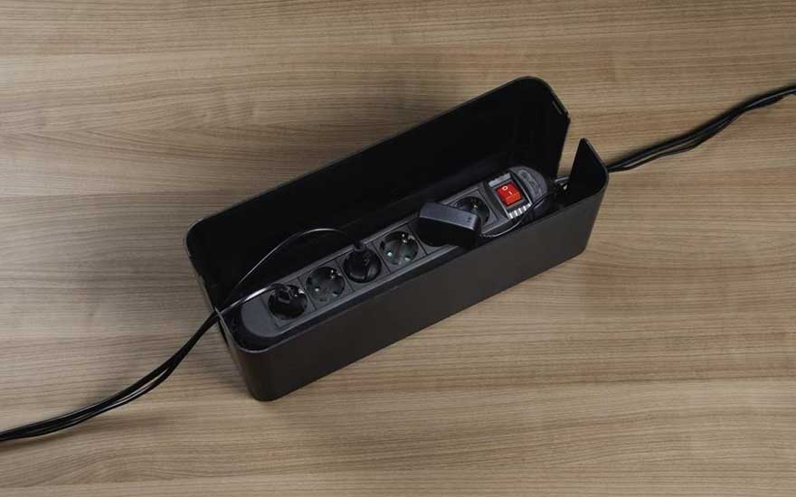 Une boîte noire ouverte sur une table en bois contient une multiprise noire avec un interrupteur rouge. Un câble noir sort de la boîte.