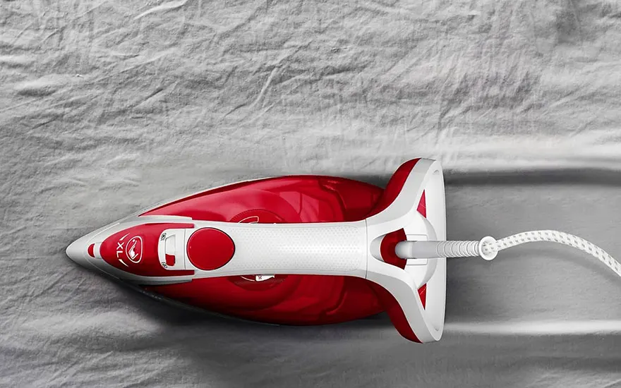 Een rood en wit strijkijzer staat op een grijze, gekreukte stof, met een snoer dat naar rechts loopt.