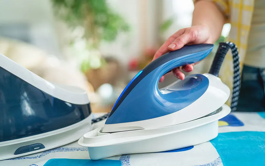 Een blauw-wit strijkijzer staat op een blauw-wit gestreepte strijkplank. Een hand houdt het strijkijzer vast.