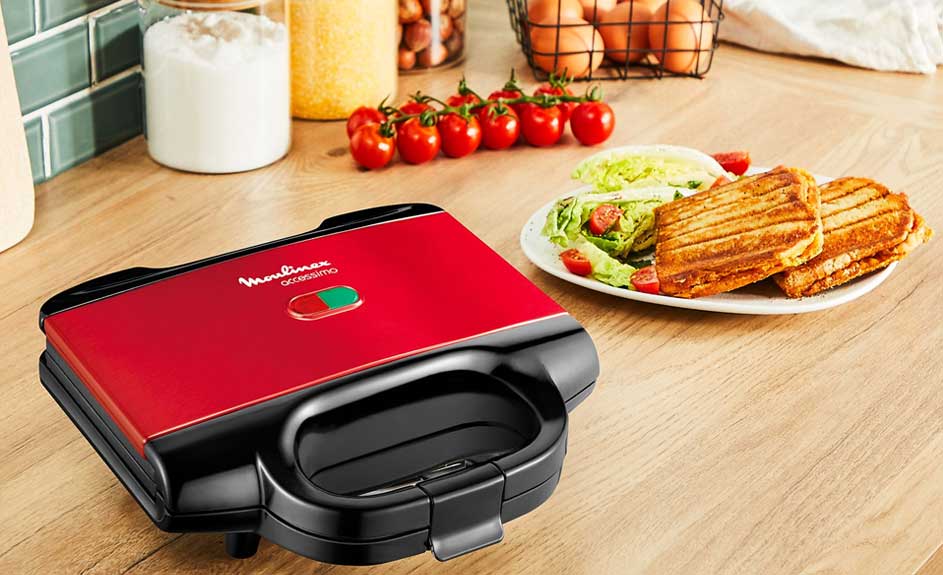 Een rode Moulinex-broodrooster staat op een houten tafel, met een bord gegrilde sandwiches en salade ernaast. Tomaten en eieren staan op de achtergrond.