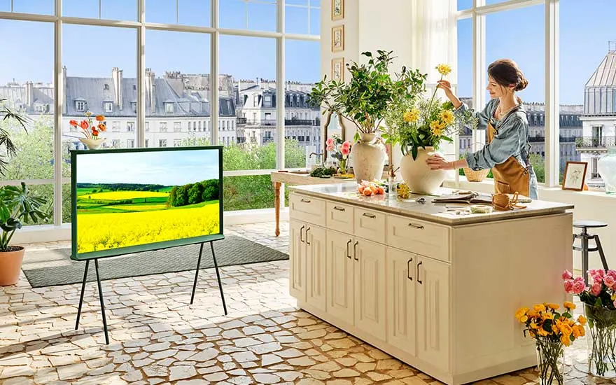 Een lichte keuken met grote ramen, een vrouw schikt bloemen aan het aanrecht en een TV toont een zonnig landschap.