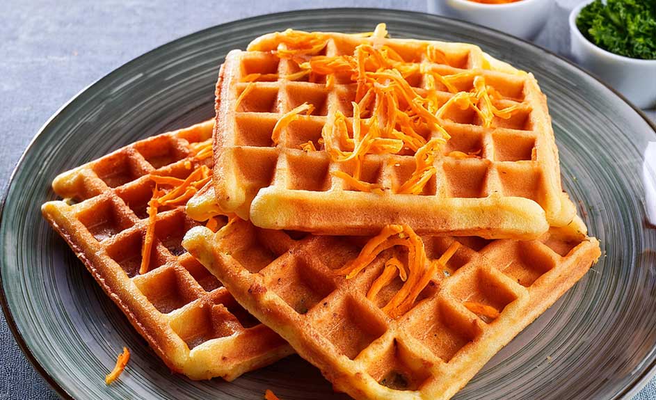Drie vierkante goudbruine wafels op een grijs bord, gegarneerd met oranje wortelreepjes. Achtergrond: kommen met groene en oranje garnering.