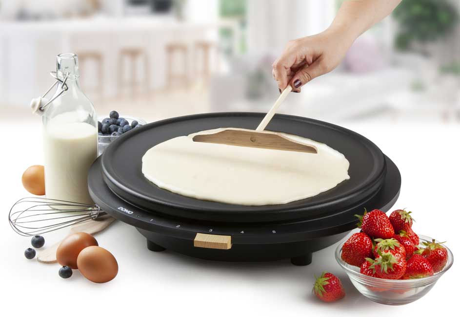 Pannenkoek op bakplaat. Melk, eieren en blauwe bessen links; aardbeien rechts. Houten spatel in de pannenkoek.