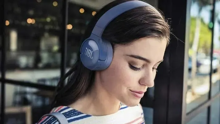 Gros plan d'un casque JBL bleu sur fond flou de fenêtre et de lumières, avec un vêtement rayé visible.