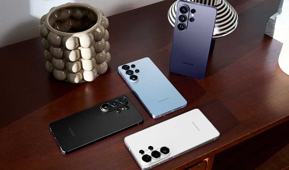 Quatre smartphones Samsung noirs, blancs, bleus et violets reposent sur une table en bois, entourés d’objets déco en céramique.