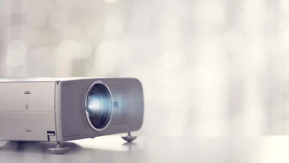 Een grijze projector staat op een tafel tegen een vage, witte achtergrond. Het licht schijnt uit de lens.