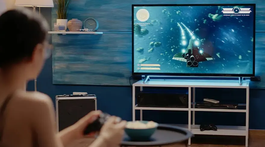 Téléviseur affichant un jeu spatial avec un vaisseau. Meuble TV blanc. Assiette sur table. Mur bleu.