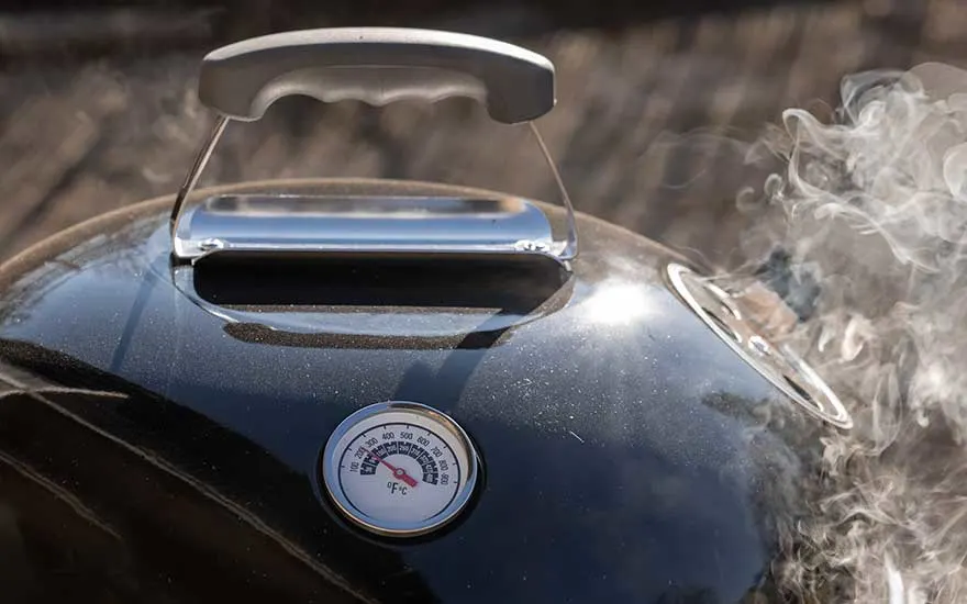 Vue rapprochée d'un barbecue noir avec thermomètre, poignée grise et fumée.