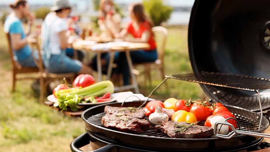 Een barbecue met vlees en groenten op een rooster. Op de achtergrond zit een groep mensen aan een tafel in een tuin.