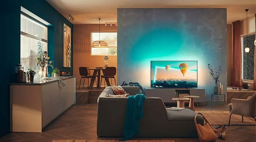 Een woonkamer met een grijze zetel, TV die een luchtballon toont, en houten vloer. Een keukenhoek is links zichtbaar.