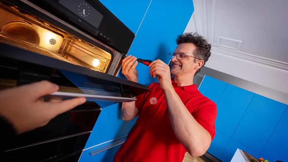 Een oven wordt gerepareerd in een blauwe keuken. Een hand houdt de deur open terwijl een schroevendraaier wordt gebruikt.