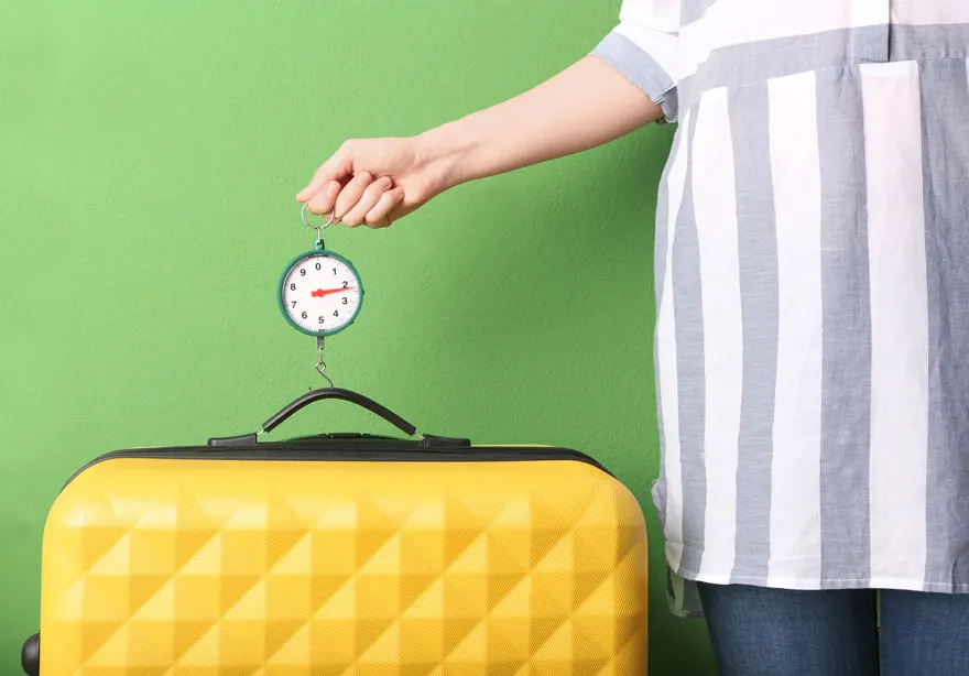 Une valise jaune est pesée avec un pèse-bagage à main sur fond vert.