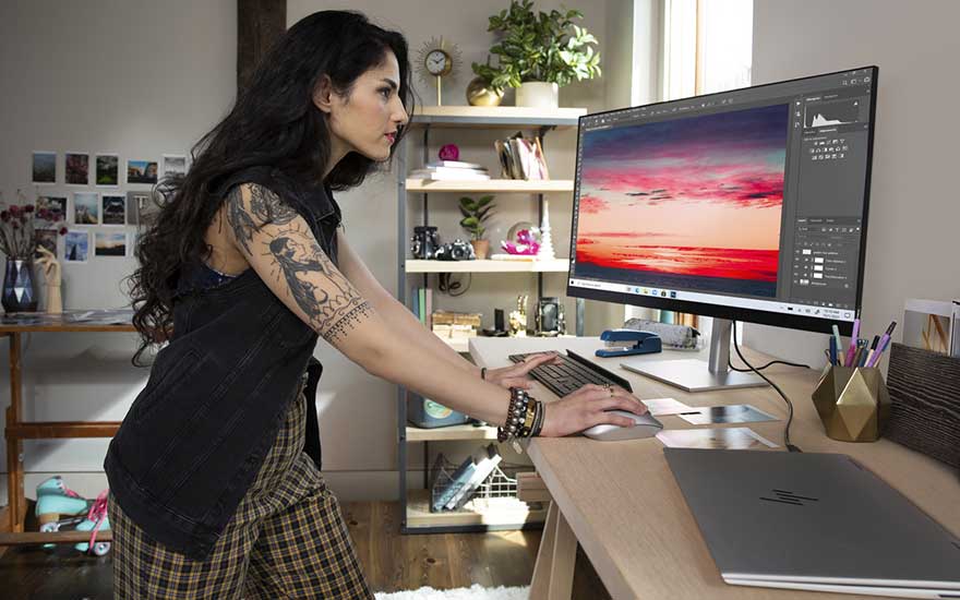 Een getatoeëerde vrouw werkt aan een bureau met toetsenbord en muis, terwijl een monitor een kleurrijke zonsondergang toont.