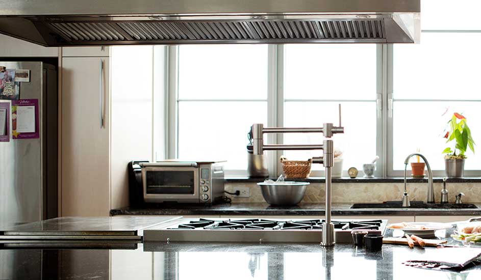 Een keuken met een kookplaat, aanrecht en roestvrijstalen afzuigkap onder een raam. Er staat een zilveren kraan.