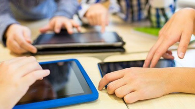 Verschillende handen bedienen tablets met blauwe en zwarte omhulsels op een lichte tafel.