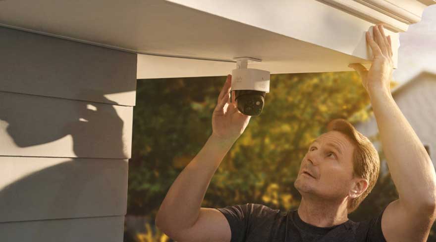 Een witte beveiligingscamera is aan een wit dak bevestigd, naast een lichtgrijze muur. Bomen en een huis zijn op de achtergrond te zien.
