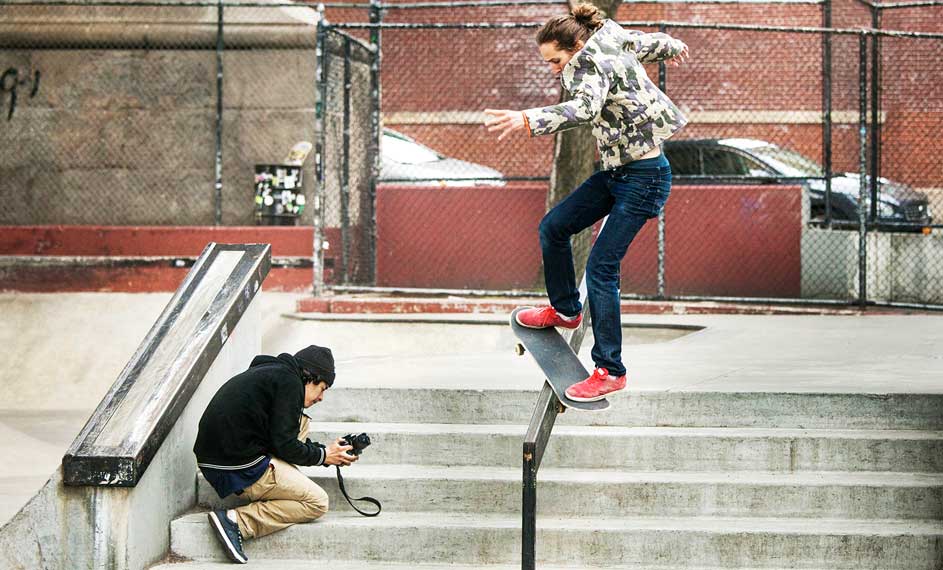 Une personne sur un skateboard sur une rampe, une autre prend une photo. Fond : clôture et voiture.