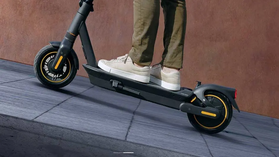 Une personne se tient sur une trottinette noire et jaune sur un trottoir gris. Un mur est visible en arrière-plan.