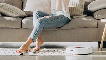 Une personne assise sur un canapé beige avec des coussins. Un robot aspirateur blanc est sur un tapis à motifs.