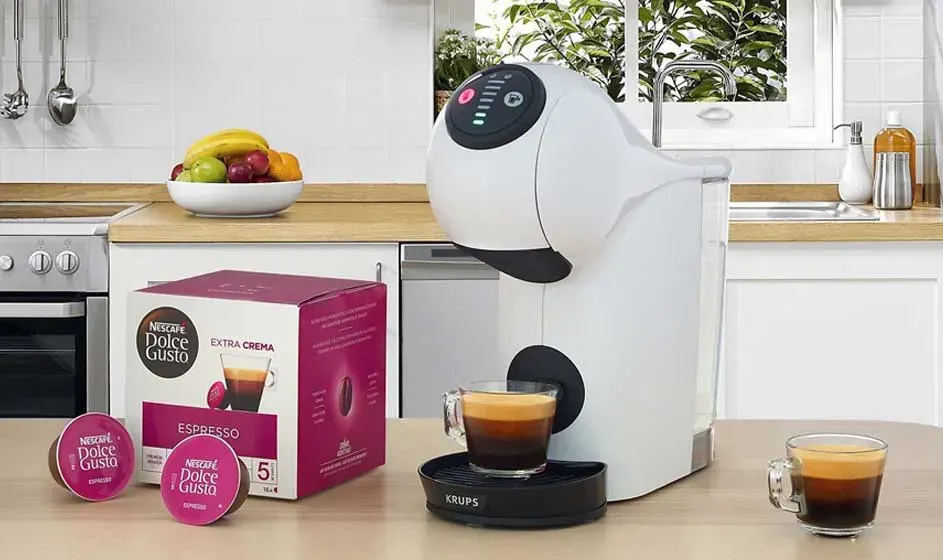 Une machine à café Dolce Gusto blanche est dans une cuisine avec capsules roses, boîte et petits verres d’espresso sur le plan.