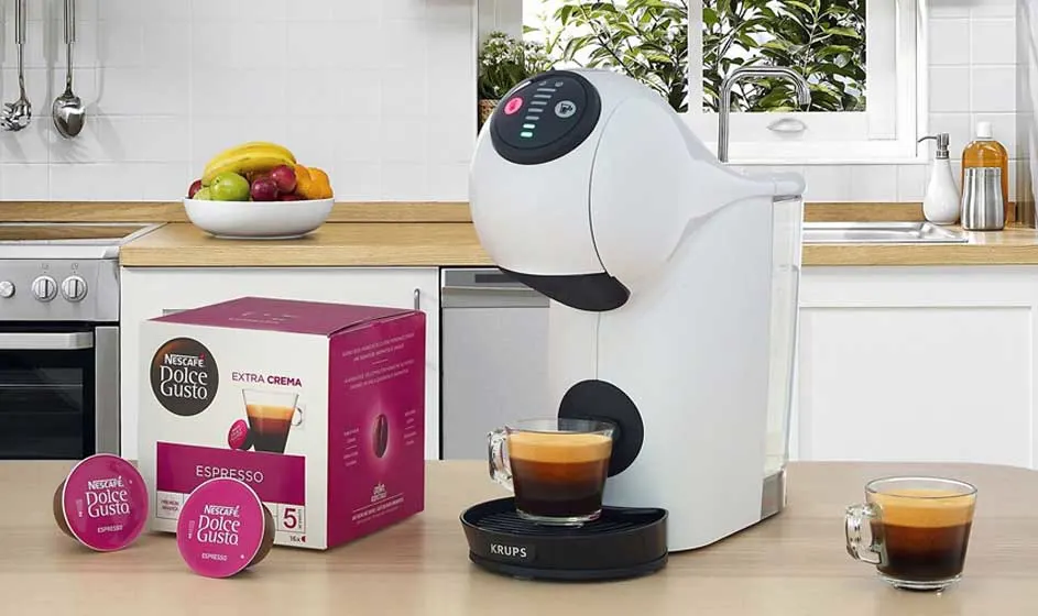 Een witte Dolce Gusto-koffiemachine staat in een keuken naast roze capsules en een doos, met kleine glazen espresso op het aanrecht.