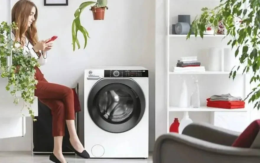 Une femme est assise avec son smartphone à côté d’une machine à laver Hoover dans une pièce lumineuse, entourée de plantes et de linge plié.
