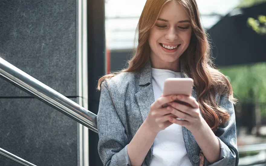 Garde-corps argenté devant un mur texturé gris. Un téléphone rose est tenu par les mains. Veste grise et t-shirt blanc.