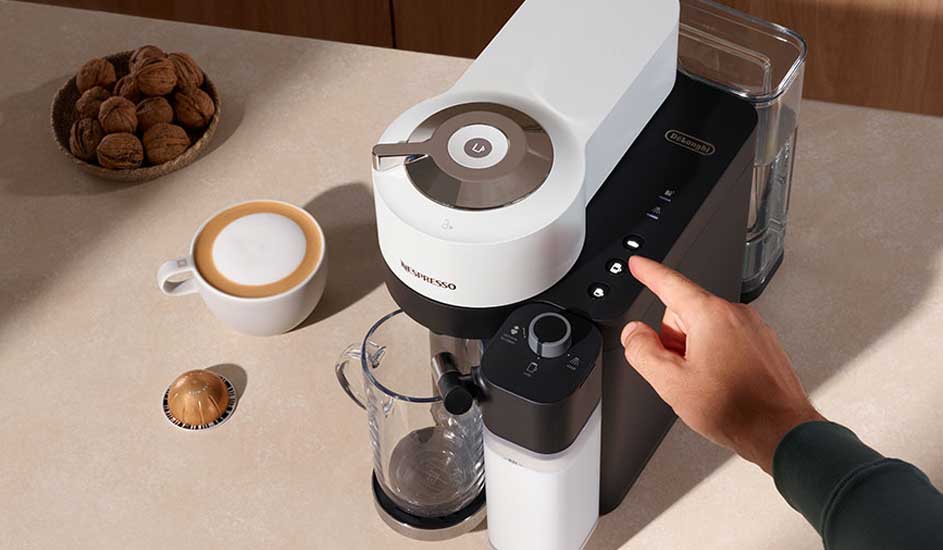 Een close-up van een Nespresso-machine, een kopje koffie, walnoten en een hand die een knop bedient.