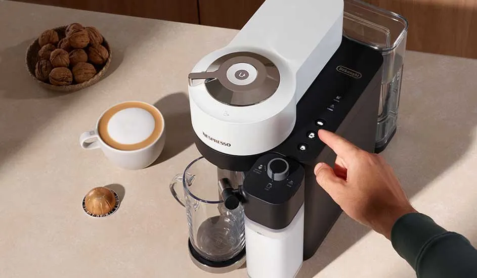 Een close-up van een Nespresso-machine, een kopje koffie, walnoten en een hand die een knop bedient.