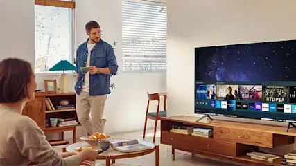Woonkamer met tv op houten kast, boeken, tafel met fruit en drinken. Raam met jaloezieën.
