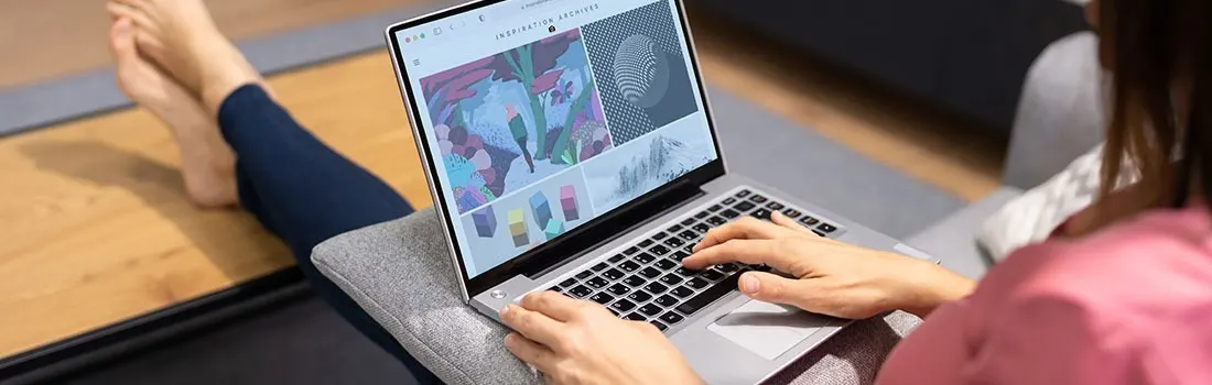 Laptop on a gray cushion displays a website with graphic designs. Hands type on the keyboard. Bare feet rest on a wooden floor.