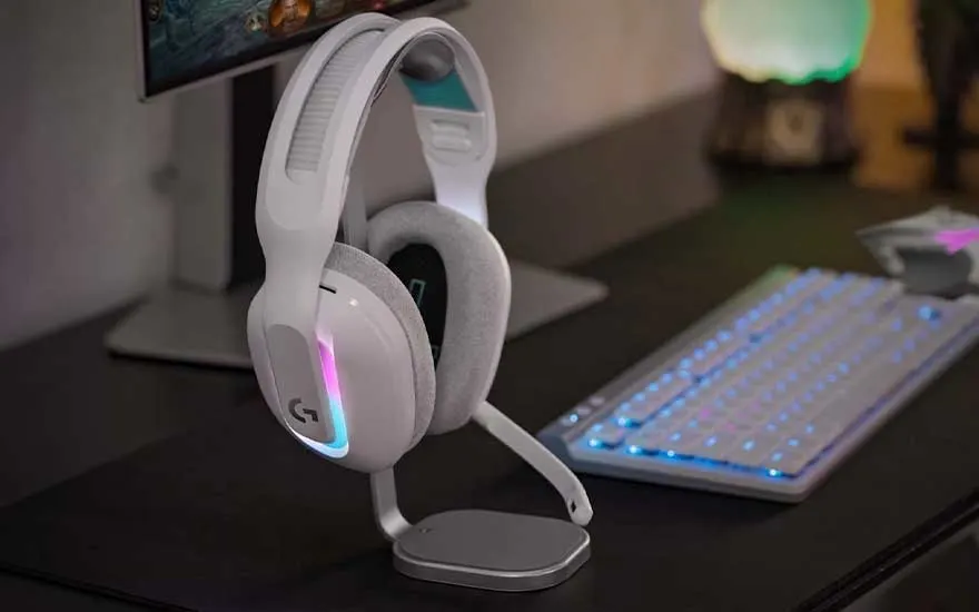Een witte gaming headset met RGB-verlichting staat op een bureau naast een verlicht toetsenbord, met een monitor en gaming-setup op de achtergrond.