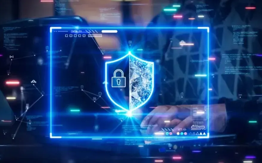 A laptop displays a glowing blue shield icon with a padlock, symbolizing data security. Digital data streams in the background.