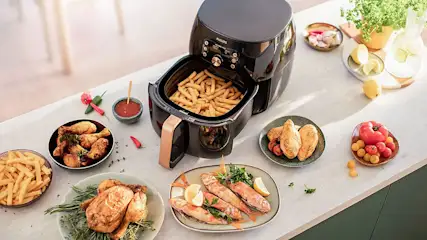 Black air fryer with fries on a table with chicken, fish, and tomatoes.