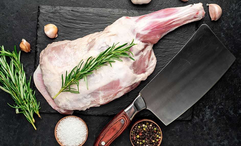 Sur une ardoise noire, une gigot d'agneau cru est décorée de romarin, à côté d'un hachoir, d'ail, de sel et de poivre.