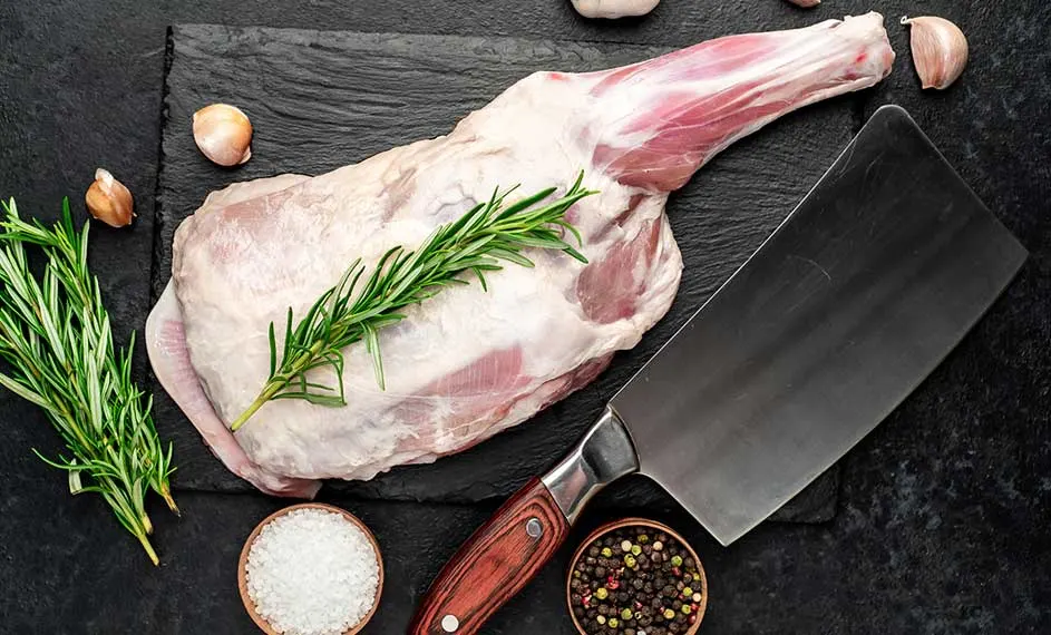 Sur une ardoise noire, une gigot d'agneau cru est décorée de romarin, à côté d'un hachoir, d'ail, de sel et de poivre.